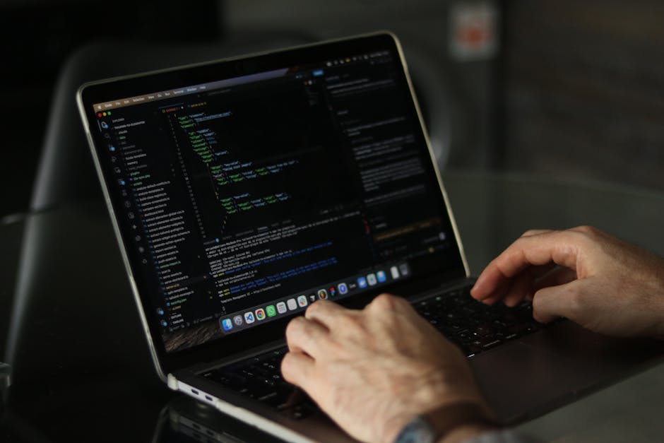 A person working on a laptop displays a dark-themed coding interface with lines of colorful code on the screen. The individual is using both hands to navigate or type, seated at a desk or table. The b